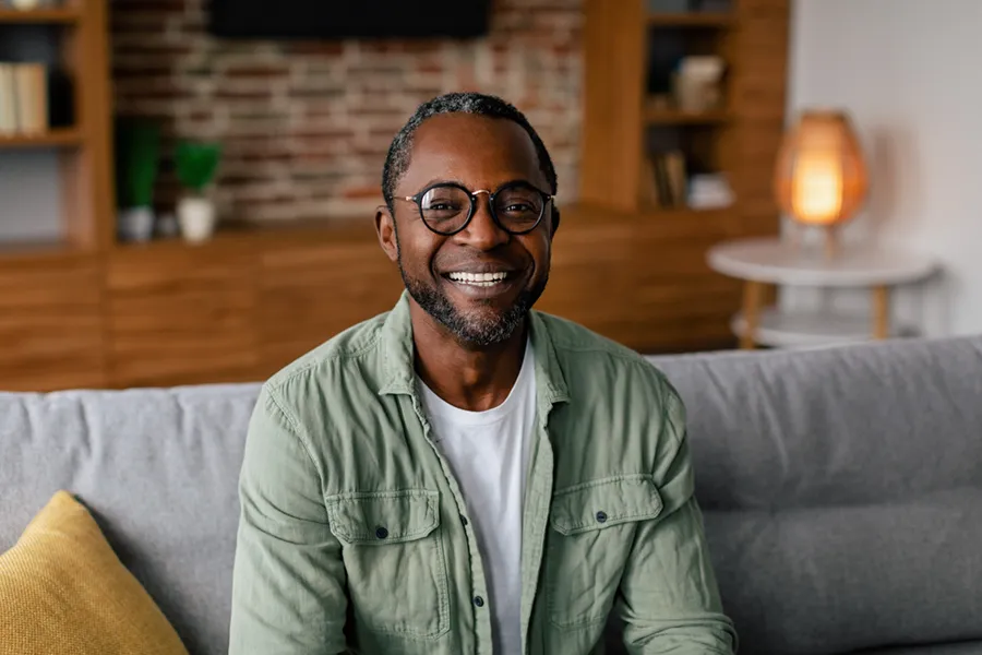 Andropause-Doctor A middle aged black man in a green button up shirt sits on his couch smiling, happy with his treatment for andropause and low testosterone from Vitality Medical in Seattle.