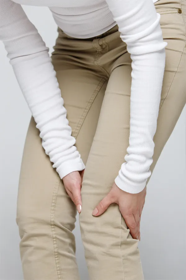 Arthritis-Treatment A woman wearing a white long-sleeve shirt and tan pants, clutching her knee, suffering from arthritis. Get arthritis treatment from Vitality Medical in Seattle.