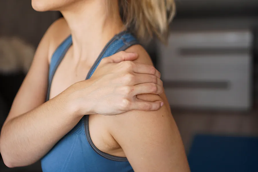 A close-up view of a woman in a blue tank top, gripping her own shoulder with a look of discomfort. Receive expert Frozen Shoulder treatment by Vitality Medical located in Seattle.