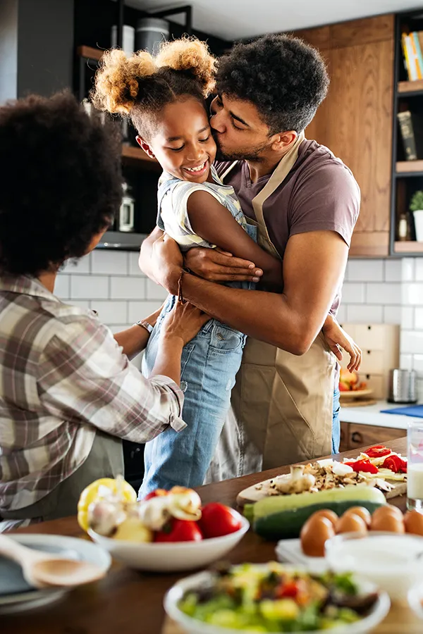 Gut-Health-Treatment A man holds his daughter and kisses her cheek as the family makes a healthy dinner in the kitchen. Get help for digestive issues and gut health from Vitality Medical in Seattle.