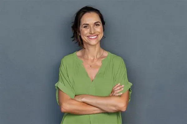 Hormones-for-Women-Doctor A woman in a green shirt stands smiling against a gray-blue wall, pleased with her hormone therapy from Vitality Medical in Seattle.