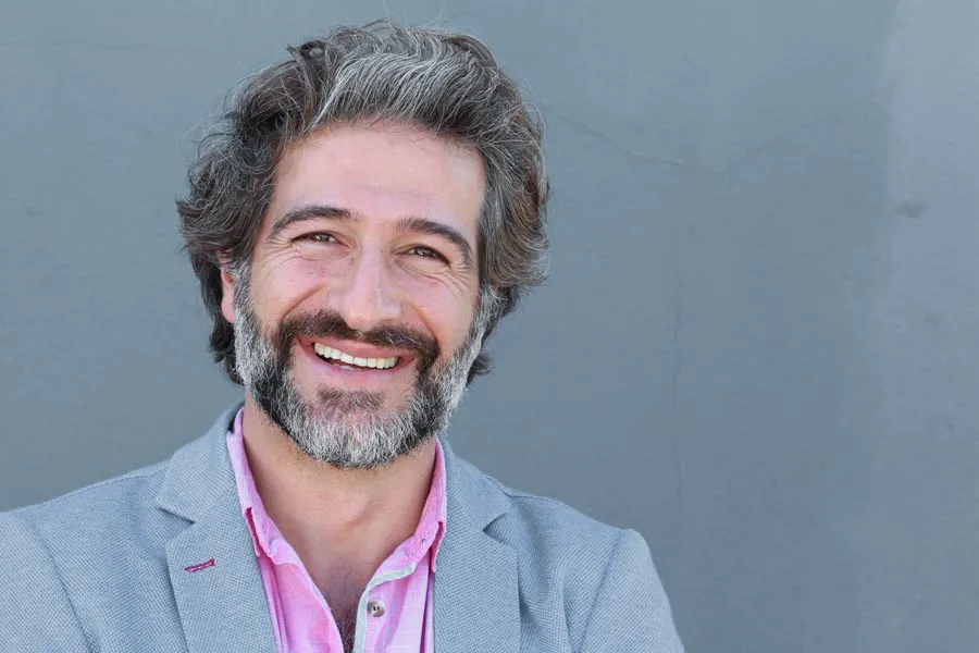 Hypogonadism-Clinic A smiling man with salt and pepper hair and beard wearing a pink shirt and gray blazer stands agains a gray wall. Get hypogonadism treatment from Vitality Medical in Seattle.
