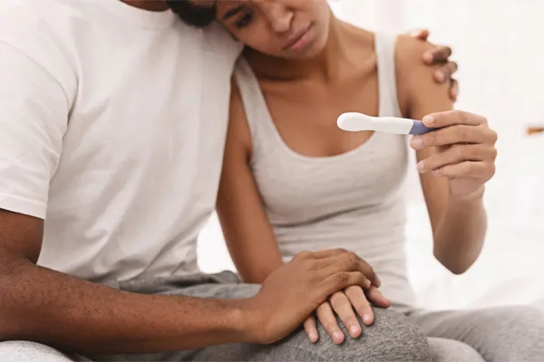 A black couple sits on a bed, disappointed with the negative results of a pregnancy test. Get treatment for infertility from Vitality Medical in Seattle.