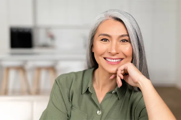 An asian woman with long gray hair and a green shirt sitting and smiling on her couch, happy with her menopause care from [NAME} in Seattle.