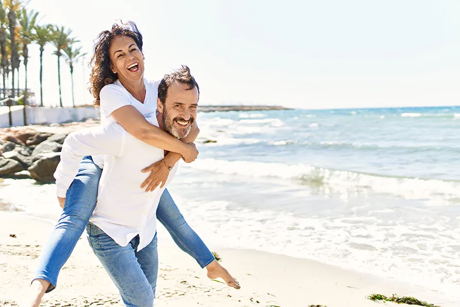 A woman piggybacks on her husband as the middle-aged couple have fun at the beach. Get NAD Therapy from Vitality Medical in Seattle.