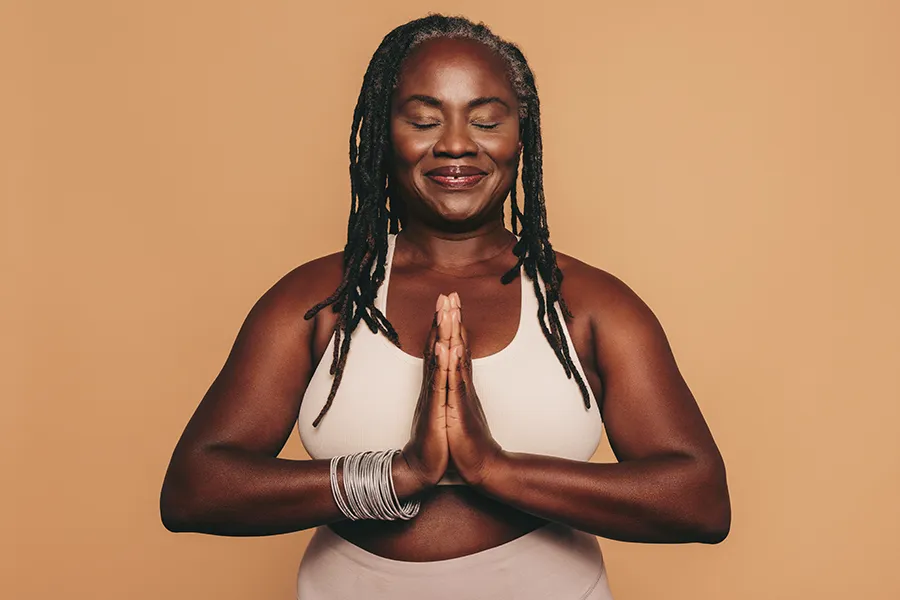 Obesity-Clinic An older African American woman confidently posing against a warm background smiling with hands in a prayer pose. Vitality Medical treats obesity using functional medicine in Seattle.