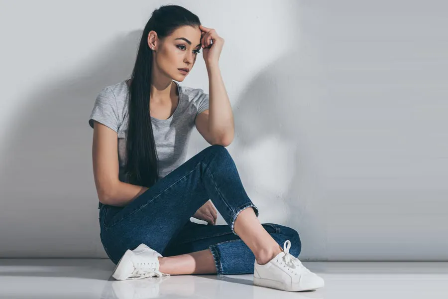PCOS-Doctor A young brunette woman in a gray t-shirt and jeans sits against a gray wall with her head resting on her hand and a distressed expression. Get help for Polycystic Ovarian Syndrome (PCOS) from Vitality Medical in Seattle.