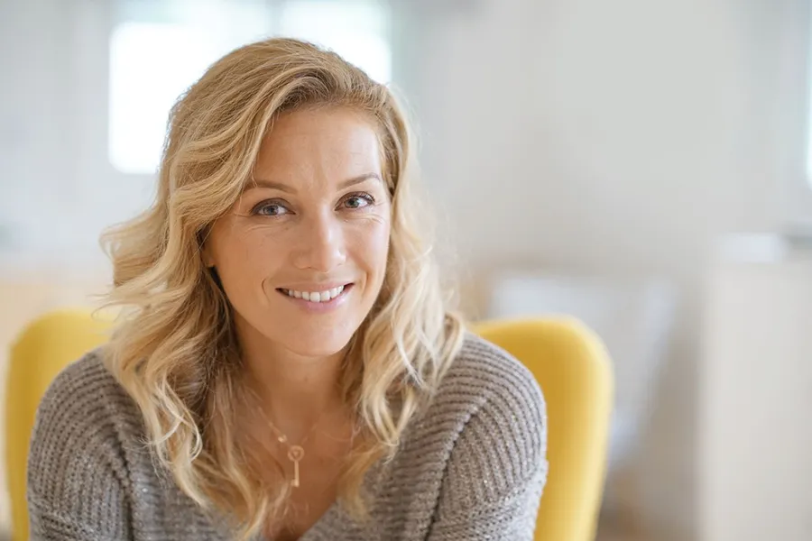 A blonde woman wearing a a gray sweater sits inside in a yellow chair, smiling and happy with her perimenopause treatment from Vitality Medical in Seattle.