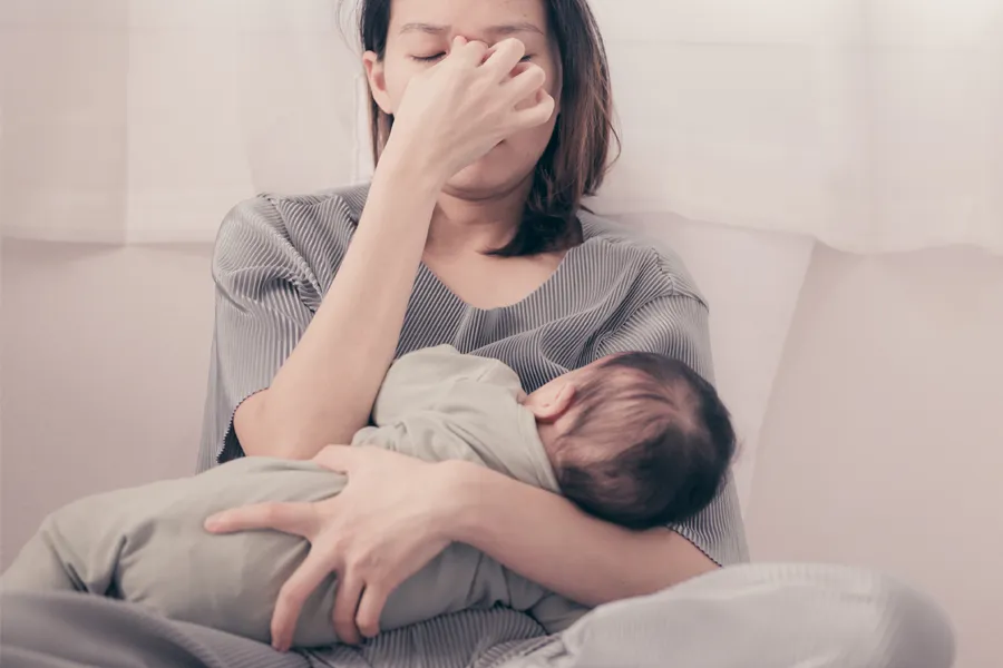 Postpartum-Hormone-Imbalance-Clinic A woman nursing an infant on couch rubs her eyes with exhaustion. Get treatment for postpartum hormone imbalance from Vitality Medical in Seattle.