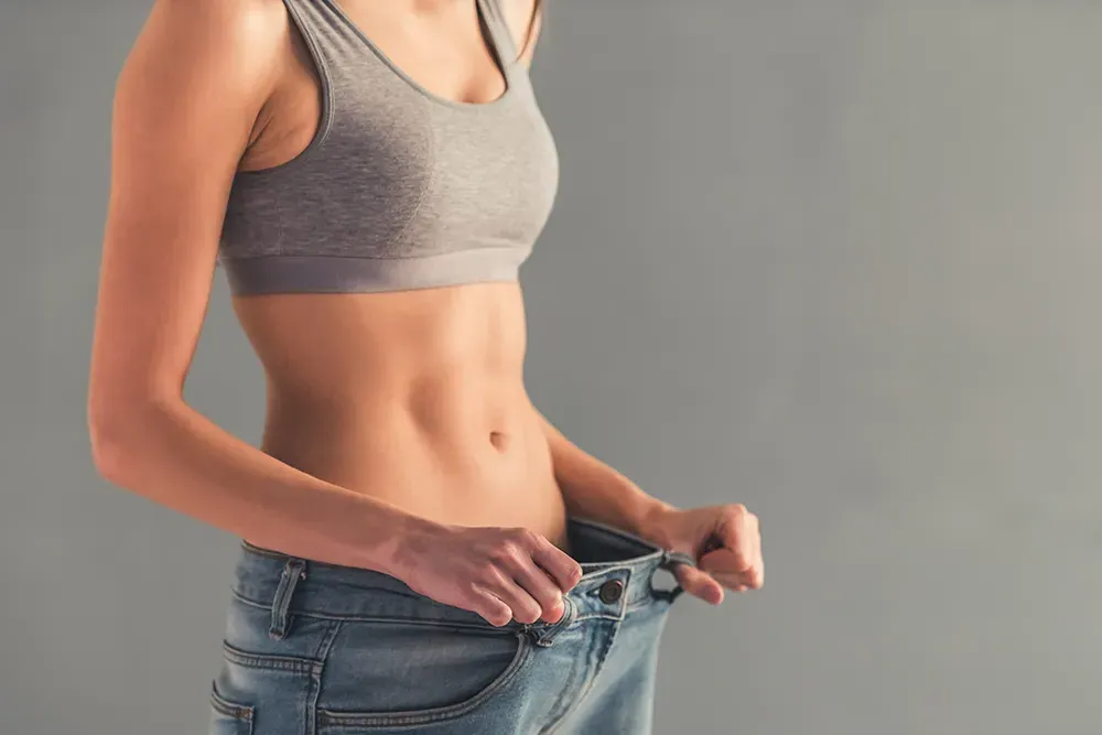 A woman pulls too-big jeans away from her waist to show weight loss. Schedule Tesamorelin peptide treatment from Vitality Medical in Seattle.