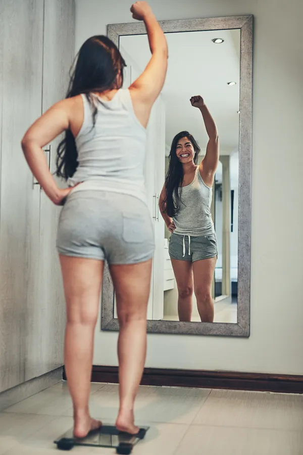 Tirzepatide-Treatment A woman in workout clothes stands on a scale looking in the mirror and pumping her hands in celebration of her successful Tirzepatide treatment from Vitality Medical in Seattle.